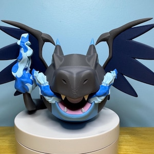May include: A 3D-printed dragon figurine in shades of gray, blue, and white. The dragon has large, dark blue wings, blue accents, and a light blue underbelly. It is posed on a white circular base.