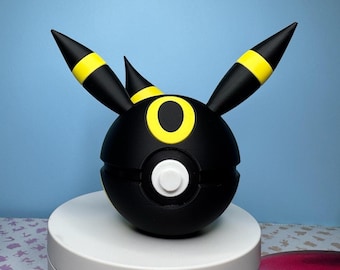 May include: A black and yellow spherical sculpture with pointed ears and a white button. The sculpture has a yellow ring around the center and yellow bands on the ears. The object is sitting on a white circular platform.