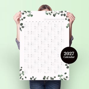 May include: A white 2027 wall calendar featuring a grid layout for the year with the text "TWO THOUSAND AND TWENTY-SEVEN" at the top. The design is decorated with green eucalyptus leaf illustrations along the top and bottom edges.