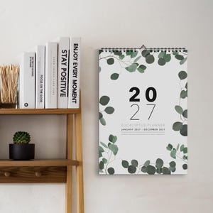 May include: A wall calendar for the year 2027 titled Eucalyptus Planner, featuring a white background with green eucalyptus leaf illustrations framing the text. The calendar includes the dates January 2027 through December 2027 with black wire binding.