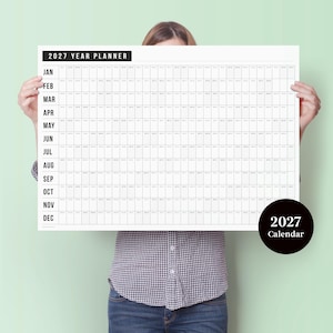 May include: A large white 2027 year planner with a black border and grid layout. The text "2027 YEAR PLANNER" is at the top, with months listed vertically. A black circle with "2027 Calendar" is in the bottom right.
