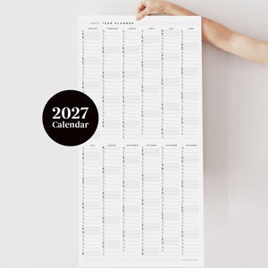 May include: A large, vertical 2027 year planner with a minimalist design. The calendar is white with black text and a black circle with "2027 Calendar" printed on it. The planner is held up by a person's hand.