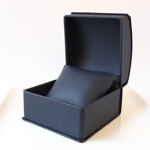 May include: A dark blue, rectangular watch box with a soft, cushioned interior. The box is open, revealing the empty cushion.