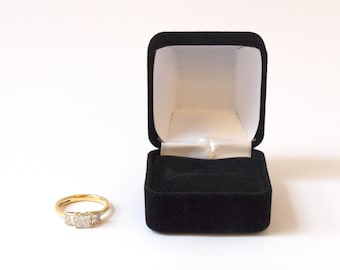 May include: A gold ring with three square-shaped diamonds set in a row. The ring is in a black velvet ring box.
