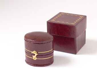 May include: Two burgundy leather ring boxes. The smaller box is round with a gold latch and two gold bands. The larger box is square with a gold decorative border.