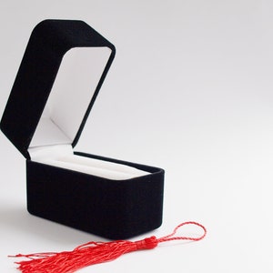 Könnte beinhalten: Eine leere schwarze Samt-Ringbox mit einem roten Quasten.