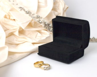 May include: A black velvet ring box with two gold wedding bands inside. The box is open and sitting on a white fabric background.