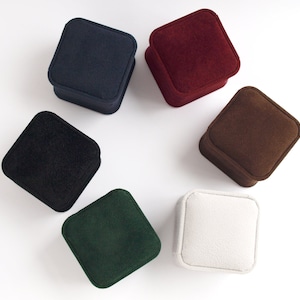 May include: Six square jewellery boxes with a soft velvet texture arranged in a circular pattern. The collection includes boxes in navy blue, deep red, dark brown, black, forest green, and off-white, each featuring rounded corners and a lid.