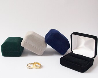 May include: Four velvet ring boxes in green, beige, blue, and black. The black box is open and has a white interior. Two gold rings are in front of the boxes.