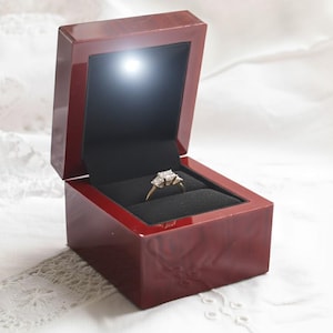 Könnte beinhalten: Eine rote, rechteckige Schmuckschatulle mit glänzendem Finish, geöffnet, um einen Ring mit drei quadratischen Steinen zu enthüllen. Der Innenraum ist schwarz, und ein kleines Licht beleuchtet den Ring. Die Schatulle steht auf einer weißen, strukturierten Oberfläche.