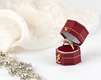 May include: A red ring box with a gold trim is open, revealing a gold ring with a large diamond and two smaller diamonds on either side. The ring is sitting on a red velvet cushion.