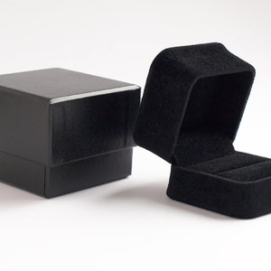 Könnte beinhalten: Eine schwarze Ringbox mit einem Samt-Innenfutter. Der Deckel ist geöffnet und die Box steht auf einer weißen Oberfläche.