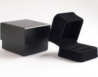 May include: A black square jewelry box with a textured exterior finish sits next to an open black velvet ring box. The open box features a soft, plush interior lining with a central slot designed to hold a single ring securely in place.