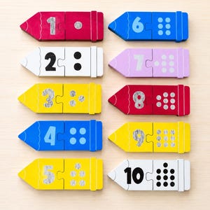 Peut inclure: Dix puzzles en bois en forme de crayon, chacun affichant un chiffre de un à dix. Les puzzles sont de couleurs vives : rouge, bleu, jaune et blanc, avec des points représentant la valeur numérique.