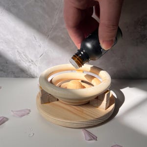 May include: A wooden essential oil diffuser with a round, multi-layered design. A hand is pouring essential oil from a dark glass bottle into the diffuser.