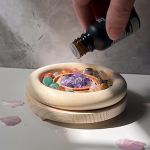 May include: A wooden diffuser with a circular base and a removable top. The top is filled with various colored gemstones, including amethyst crystals. A hand is pouring essential oil from a dark glass bottle into the diffuser.