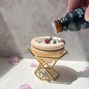 May include: A wooden bowl filled with colorful stones sits on a gold metal stand. A bottle of essential oil labeled "Better & Co" is being poured into the bowl.