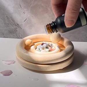 May include: A wooden diffuser with a circular design, featuring a small bowl filled with colorful stones. A hand is holding a bottle of essential oil over the bowl, ready to be added.
