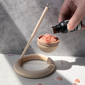 May include: A wooden diffuser with a hanging bowl filled with pink Himalayan salt crystals. A hand is pouring essential oil into the bowl.