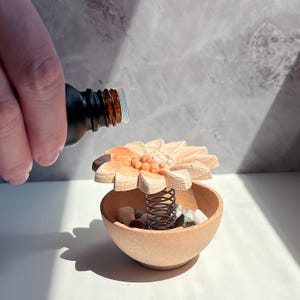 May include: A wooden flower-shaped essential oil diffuser with a brown glass bottle pouring oil into it. The diffuser has a spring and a small bowl filled with stones. The flower is light brown with orange accents.