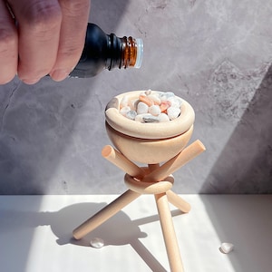 May include: A small, light-colored wooden bowl on a tripod stand, filled with small, colorful stones. A dark bottle is pouring liquid into the bowl. The bowl and stand are made of natural wood, and the stones are white, pink, and gray.