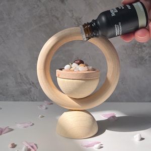 May include: A wooden diffuser with a round opening and a small bowl filled with stones. A black bottle with a dropper is being poured into the bowl. The bottle label reads "Badger & Co. Peppermint Essential Oil".