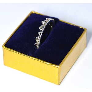 Può includere: Un anello color argento con un design a corona decorativa, presentato in una scatola dorata con interno in velluto blu. I dettagli intricati dell'anello sono evidenziati sullo sfondo scuro, mostrando il suo elegante design.