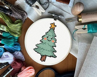 May include: A cross-stitch Christmas tree design in a white embroidery hoop, with a gold star on top. Various embroidery supplies, including thread, scissors, and needles, are arranged around the hoop on a wooden surface.