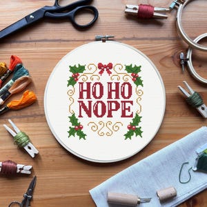 Puede incluir: Un bastidor de punto de cruz blanco con el texto "HO HO NOPE" en rojo, rodeado de acebo verde y espirales dorados. Suministros de costura como tijeras, hilo y pinzas para la ropa sobre una superficie de madera.