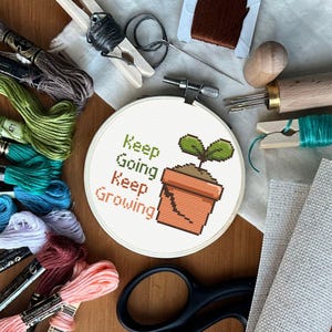 Puede incluir: Un bastidor de bordado de punto de cruz con el texto "Keep Going Keep Growing" y un diseño de planta en maceta. La imagen incluye hilo de bordar, tijeras, agujas y otros suministros de artesanía, todo dispuesto sobre una superficie de madera.