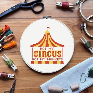 Puede incluir: Un bastidor de punto de cruz con un diseño de carpa de circo roja y amarilla y el texto "NOT MY CIRCUS NOT MY MONKEYS". La imagen incluye suministros de bordado como tijeras, hilo y pinzas para la ropa, todo sobre una superficie de madera.