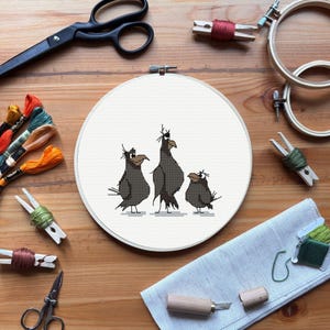 May include: A cross-stitch embroidery hoop featuring three stylized black birds with expressive faces. The artwork is surrounded by embroidery supplies, including scissors, thread, and wooden hoops, all arranged on a wooden surface. The birds are the focal point.
