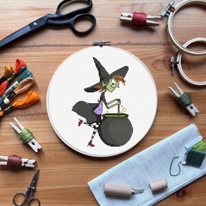 May include: A cross-stitch embroidery hoop featuring a cartoon witch stirring a cauldron. The witch wears a black hat, dress, and striped stockings. The scene is surrounded by embroidery supplies, including scissors, thread, and wooden hoops, on a wooden surface.
