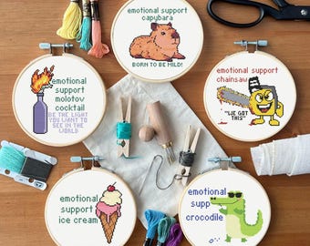 May include: Five embroidery hoops with different designs. The hoops are on a wooden surface with embroidery floss, a needle, and other embroidery supplies. The designs include a capybara, a chainsaw, a molotov cocktail, ice cream, and a crocodile. The text on the hoops reads "emotional support capybara", "emotional support chainsaw", "emotional support molotov cocktail", "emotional support ice cream", and "emotional support crocodile".