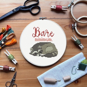 Puede incluir: Un bastidor de bordado con un diseño de mapache y el texto "Bare minimum". La imagen incluye hilo de bordar, tijeras, pinzas de madera y otros suministros de manualidades sobre una superficie de madera, ideal para artesanos.