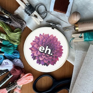 May include: A close-up of a purple flower cross-stitch embroidery hoop with the word "eh." in white text. The hoop is surrounded by embroidery floss, scissors, and other embroidery supplies.