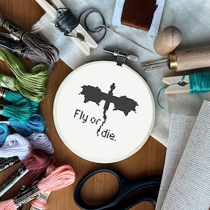 Puede incluir: Un bastidor de bordado de punto de cruz con un diseño de libélula negra y el texto "Fly or die". Varios hilos de bordar de colores, tijeras, agujas y otros suministros de artesanía están dispuestos alrededor del bastidor.