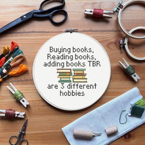 May include: A cross-stitch hoop with the text "Buying books, Reading books, adding books TBR are 3 different hobbies" and a stack of books. Embroidery supplies, scissors, and thread are arranged around the hoop on a wooden surface.