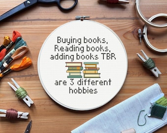 Op de afbeelding: Een borduurring met de tekst "Buying books, Reading books, adding books TBR are 3 different hobbies" en een stapel boeken. Borduurbenodigdheden, scharen en garen zijn rond de ring op een houten oppervlak geplaatst.