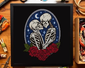 May include: Framed cross-stitch art featuring two skeletons embracing, set against a blue night sky with a crescent moon and stars. Red roses are at the bottom. The frame is black. The artwork is on a wooden surface.