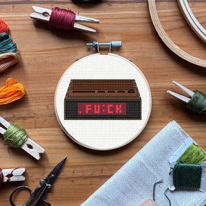 May include: A cross-stitch embroidery hoop featuring a brown retro-style digital clock design with the word "FUCK" in red. The embroidery is set against a cream-colored fabric. Various craft supplies, including thread and scissors, surround the hoop.