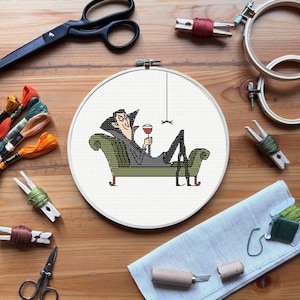 May include: A cross-stitch embroidery hoop featuring a cartoon Dracula reclining on a green chaise lounge, holding a glass of red wine. The scene is surrounded by embroidery supplies, including scissors, thread, and wooden hoops, on a wooden surface.