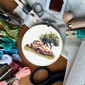 Puede incluir: Un bastidor de bordado con un diseño de punto de cruz de un coche antiguo bajo un árbol. La imagen incluye hilos de bordar, tijeras, agujas y otros artículos de artesanía, lo que sugiere un proyecto de manualidades.