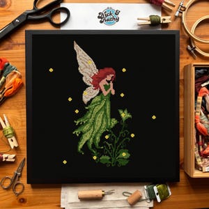 May include: A framed cross-stitch depicting a fairy with red hair and a green dress, set against a black backdrop. The fairy has detailed wings. Yellow star accents are scattered around the fairy. The frame is black.