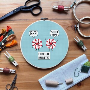 May include: A cross-stitch hoop with a light blue background features two cartoon peppermint candies with the text "6-7!" and "SHUT UP!" in speech bubbles. The text "ARGUE MINTS" is below the candies. Embroidery supplies surround the hoop.