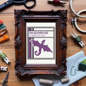 May include: A framed cross-stitch featuring a purple scorpion tail design with the text "ANDARNA SCORPION TAIL". The frame is ornate, dark brown, and sits on a wooden surface. Crafting supplies, including scissors and embroidery floss, surround the frame.
