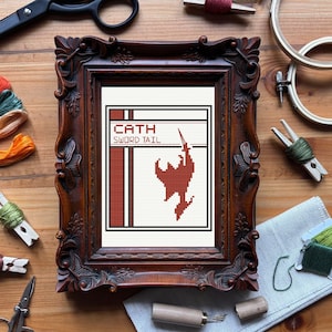 Puede incluir: Un bordado de punto de cruz enmarcado con el texto "CATH SWORD TAIL" y una silueta roja de una figura con una espada. El marco ornamentado, marrón oscuro, está sobre una superficie de madera. Suministros de bordado, incluyendo tijeras e hilo, rodean el marco.