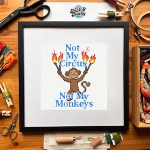 Puede incluir: Arte de punto de cruz enmarcado en un marco negro. La obra presenta un mono marrón sosteniendo antorchas con llamas, y el texto "Not My Circus, Not My Monkeys" en azul. El fondo es blanco.