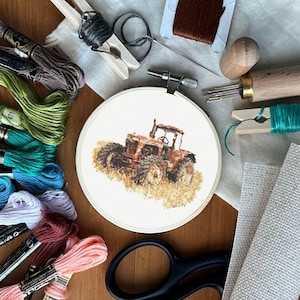 May include: A cross-stitch embroidery hoop featuring a detailed image of a vintage tractor in shades of brown and orange. The scene is surrounded by colorful embroidery floss, scissors, needles, and other craft supplies, suggesting a crafting theme.