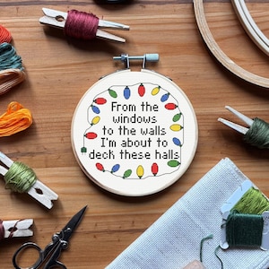 Può includere: Un telaio da ricamo a punto croce con la scritta "From the windows to the walls I'm about to deck these halls" circondata da luci natalizie colorate. Materiali per l'artigianato: filo, forbici e mollette di legno.